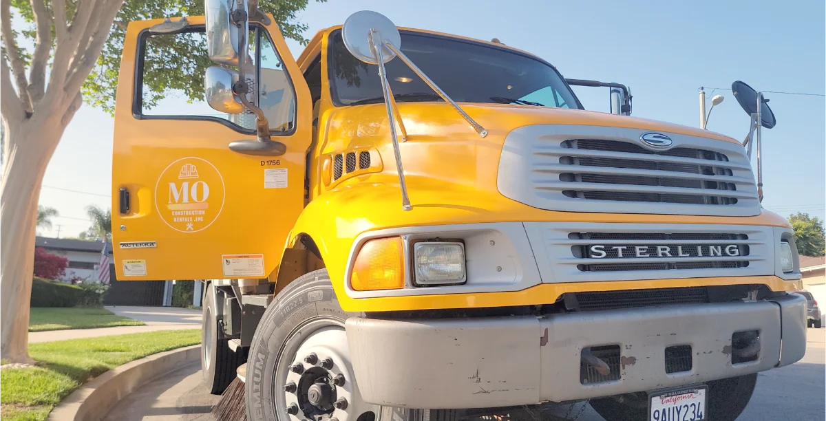 Yellow MO Construction Rentals street sweeper truck parked on a residential street, company logo on the open cab door