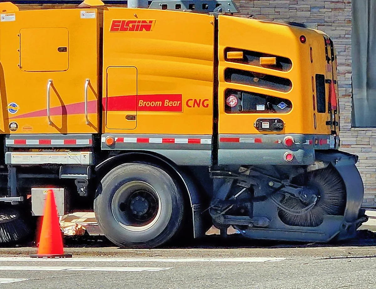 An Elgin Broom Bear CNG street sweeper working a curb, its main broom down against the pavement.